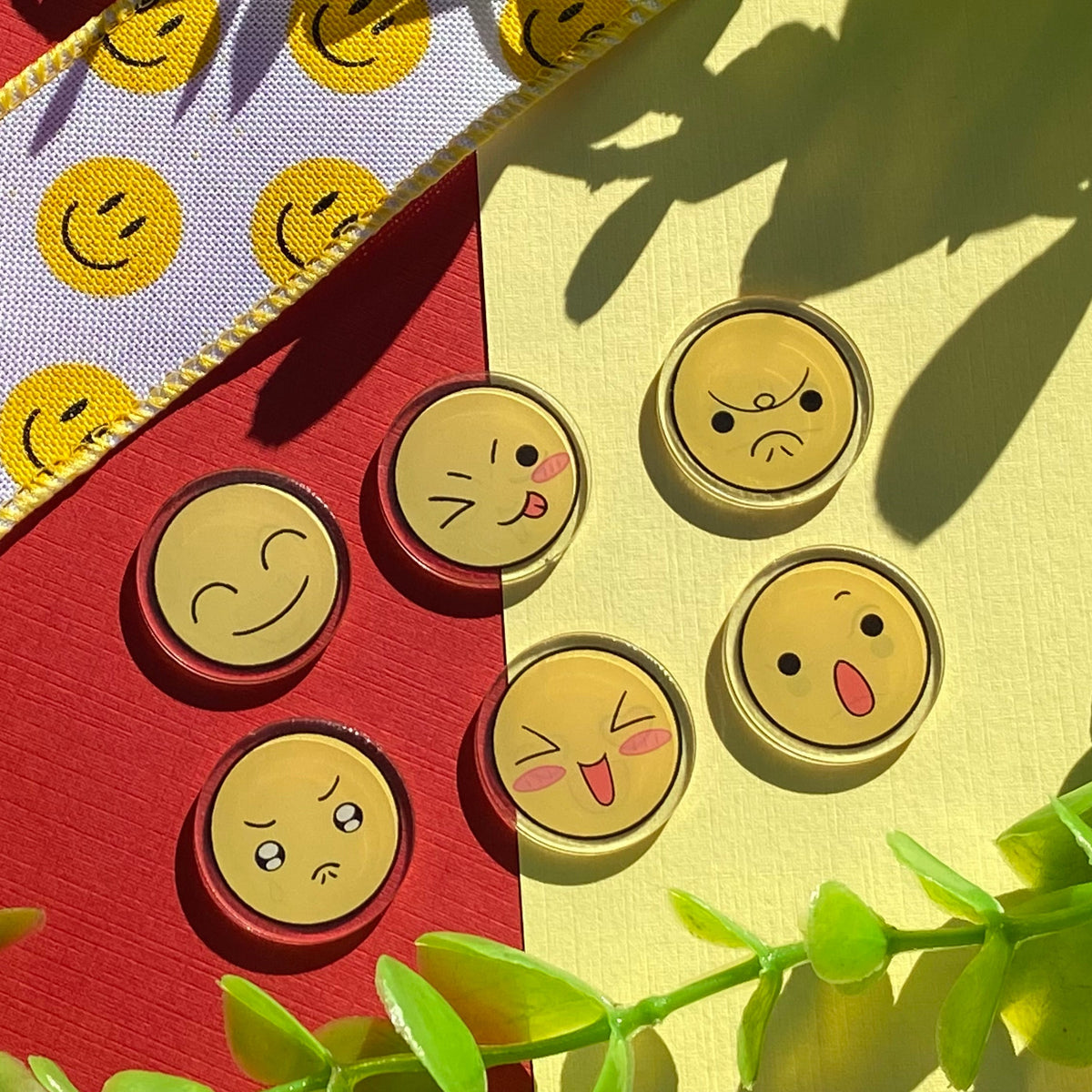 Five round  acrylic charms featuring different yellow emoji faces, arranged on a split red and pale yellow background with decorative green leaves and a patterned ribbon.