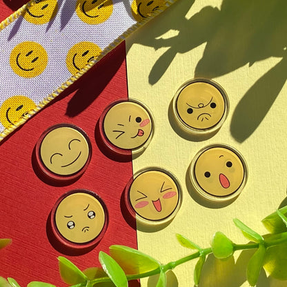 Five round  acrylic charms featuring different yellow emoji faces, arranged on a split red and pale yellow background with decorative green leaves and a patterned ribbon.